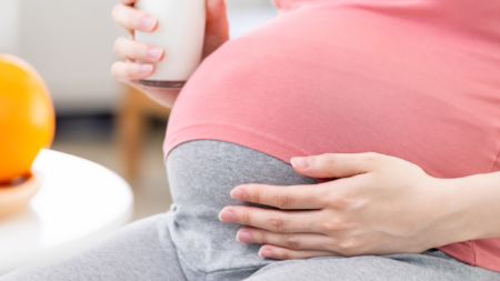 Mujer embarazada en ropa cómoda sonriendo mientras toma un batido de proteínas, en un entorno luminoso y natural